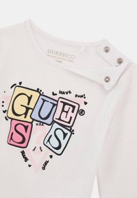 Fehér pamut póló kerek nyakkivágással és pattintós záródással. Jellemzője a színes blokkbetűk, amelyek "GUESS" feliratot alkotnak, valamint játékos grafikák.