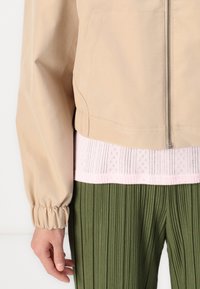 Veste beiges zippée avec des poignets élastiques, superposée à un haut texturé rose clair et un pantalon plissé vert, mettant en avant des couleurs et des matériaux distincts.