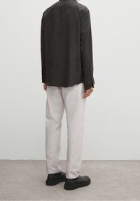 Czarna koszula z długim rękawem i mankietami zapinanymi na guziki, zestawiona z jasnymi spodniami i czarnymi butami, widziana z tyłu na jednolitym tle.