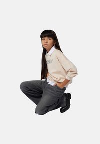 Joven arrodillado, vistiendo un sudadera beige sobre una camisa blanca, jeans grises y botas negras gruesas, contra un fondo claro y liso.