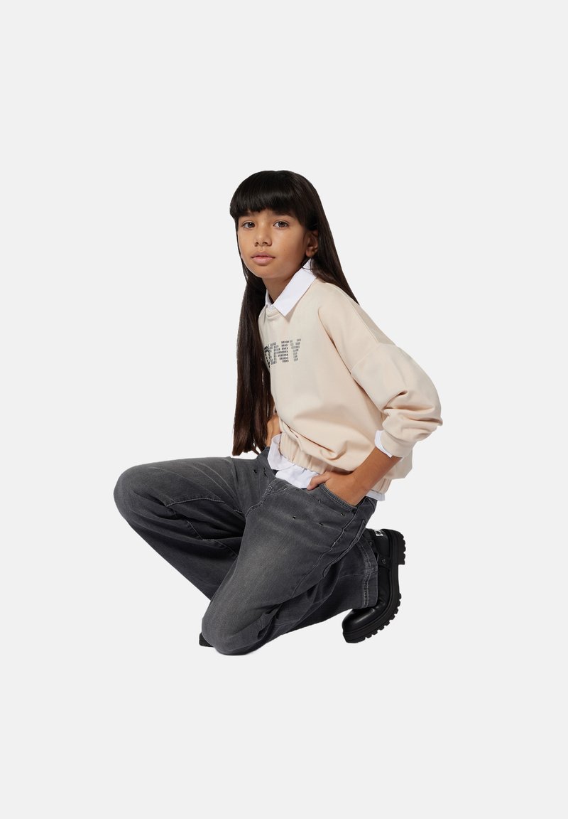 Joven arrodillado, vistiendo un sudadera beige sobre una camisa blanca, jeans grises y botas negras gruesas, contra un fondo claro y liso.