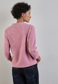 Rosa, flauschiger Strickpullover mit Dreiviertelärmeln, weicher Textur und lockerer Passform. Getragen über dunklen Jeans, mit rundem Halsausschnitt.