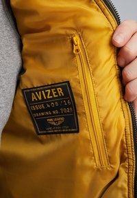 Gelbe Innenseite einer Jacke mit einer Reißverschlusstasche. Merkmale ein schwarzes Etikett mit dem Text "AVIZER" und "PME LEGEND". Weiche, glatte Textur.