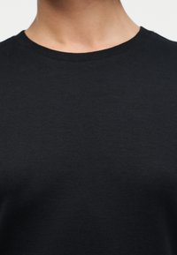 Κοντινό στον λαιμό και το πάνω μέρος του στήθους με μαύρο μονόχρωμο μπλουζάκι με στρογγυλή λαιμόκοψη σε ανοιχτό φόντο.