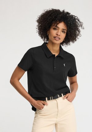 Kobieta o kręconych włosach, ubrana w czarną koszulkę polo i beżowe spodnie, stoi z rękami w kieszeniach na jednolitym tle.