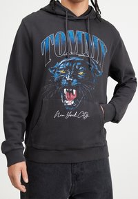 Mustahuppari, jossa on sinivalkoinen graafinen kuva karjuvasta pantterista ja teksti "TOMMY", sekä kengurupussitasku ja ribbireunat.