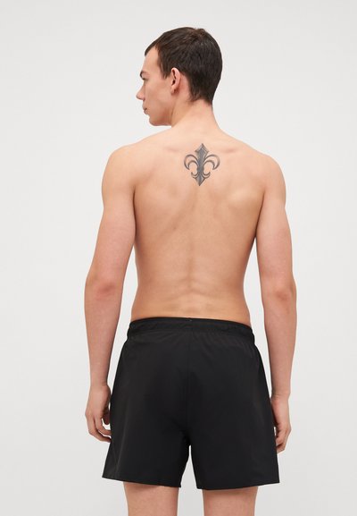Bañador negro con cinturilla elástica, textura suave y un corte holgado. Un tatuaje floral es visible en la parte posterior.