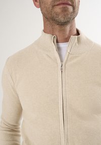 Beige gerippter Strick-Reißverschluss-Pullover mit hohem Kragen, ausgestattet mit einem glatten Reißverschluss und strukturiertem Stoff, der eine taillierte Optik bietet.
