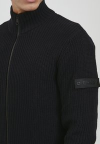 Dunkelblauer gerippter Strickzipper mit hohem Kragen und einem schwarzen Akzentpatch am linken Arm, ausgestattet mit einem eleganten Reißverschluss und strukturiertem Stoff.