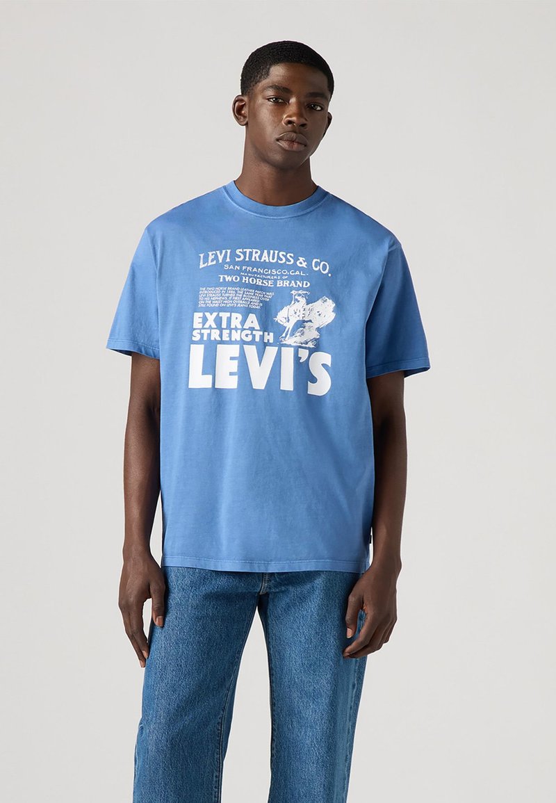 Tricou din bumbac albastru deschis cu mâneci scurte, având text și grafică imprimate în alb, cu o croială lejeră. Asortat cu blugi din denim albastru.