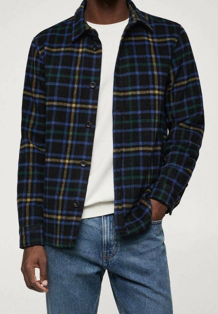 Camicia di flanella scozzese con colori nero, blu, verde e giallo; chiusura con bottoni, colletto a punta e tessuto testurizzato. Indossata sopra una camicia bianca.