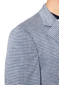 Tkanina żakietowa z wzorem houndstooth w granatowo-białym kolorze, z wyraźnym kołnierzem i teksturowanym materiałem. Zbliżenie na detal ramienia.