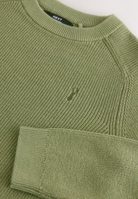 Oliivinvihreä neulepaita, jossa on ribbineulos, pyöreä kaula-aukko ja pieni kirjailtu logo rinnan lähellä. Pitkät, istuvat hihat.