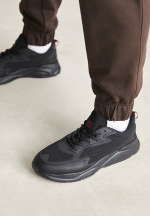 Person trägt schwarze Sportschuhe mit rotem Logo, weißen Socken und braunen Jogginghosen auf hellem Boden.