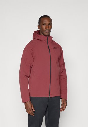 Giacca impermeabile rossa con cappuccio, chiusura a zip e tasche laterali. Presenta una texture traspirante e un logo nero sul petto.