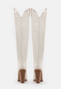 Pár vysokých bílých kožených bot s špičatými špičkami a naskládanými dřevěnými blokovými podpatky, zobrazený zezadu na bílém pozadí.