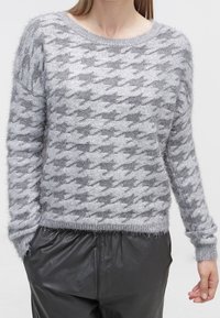 Donna che indossa un maglione grigio peloso con un grande motivo a pied-de-poule e pantaloni di pelle nera, in piedi con le mani nelle tasche.