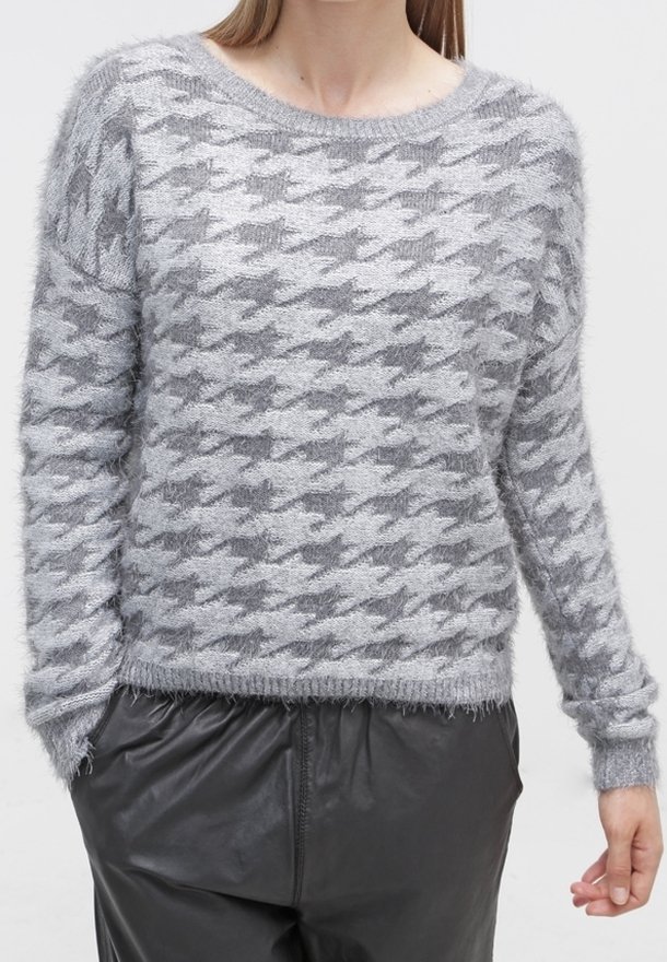 Donna che indossa un maglione grigio peloso con un grande motivo a pied-de-poule e pantaloni di pelle nera, in piedi con le mani nelle tasche.