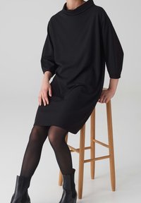 Personne assise sur un tabouret en bois portant une robe noire mi-genoux avec des manches trois-quarts, des collants noirs et des bottines noires.