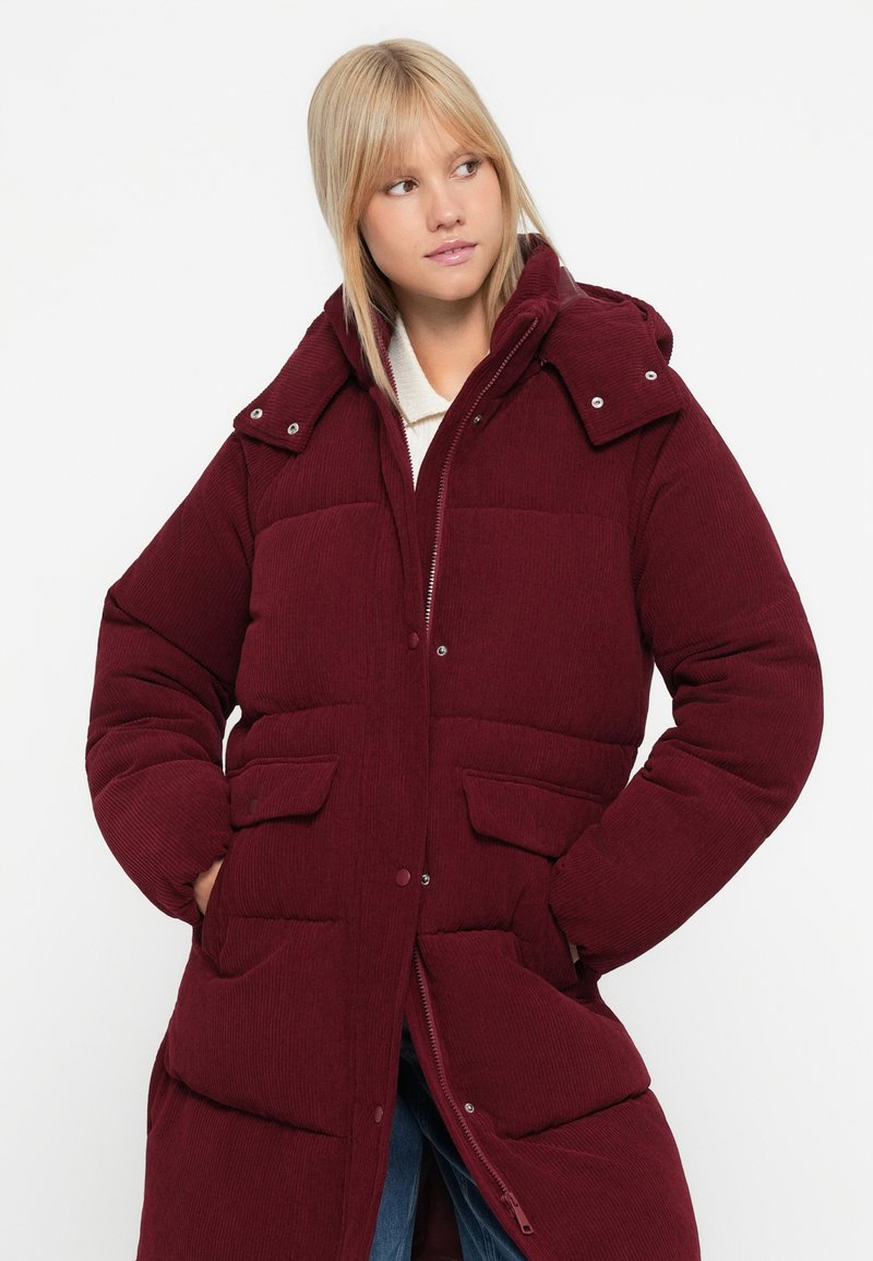 Abrigo acolchado de pana burdeos con cierre de cremallera, capucha y dos bolsillos delanteros. Presenta una superficie texturizada y un diseño acanalado.