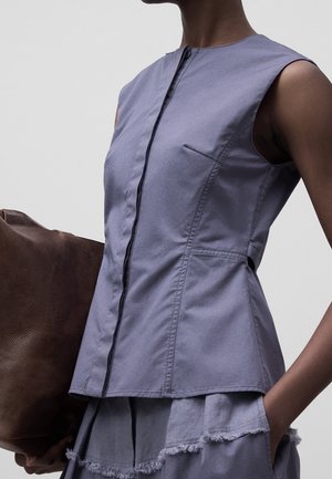 Personne portant un haut sans manches violet clair avec des détails de couture à l'avant, tenant un grand sac en cuir marron et portant une jupe violette assortie.