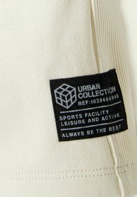 Tissu crème avec texture côtelée arborant une étiquette noire portant la mention "URBAN COLLECTION," détaillant des attributs sportifs et de loisirs.