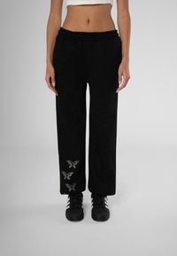 Black sweatpants with an elastic waistband, wide-leg design, featuring silver butterfly embellishments on the lower leg. Paired with black shoes.