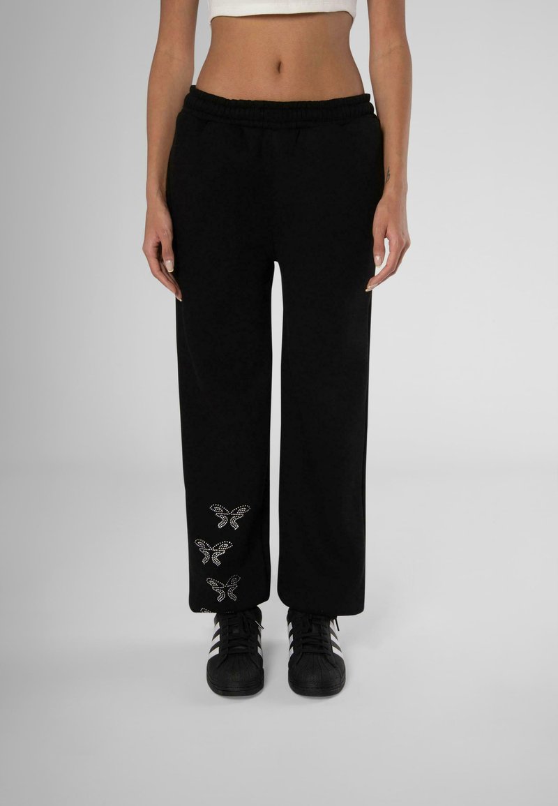 Black sweatpants with an elastic waistband, wide-leg design, featuring silver butterfly embellishments on the lower leg. Paired with black shoes.