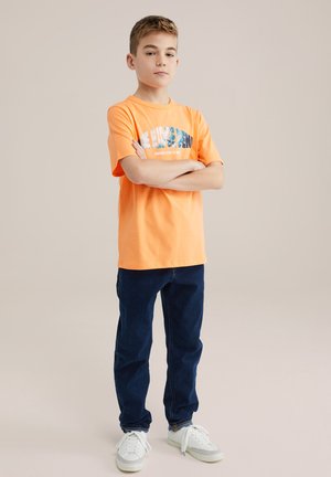 Kortarmig oranje T-shirt met bedrukte tekst, gecombineerd met donkerblauwe jeans en witte sneakers. Eenvoudig ontwerp, casual pasvorm, katoenen materiaal.