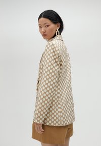 Beige mönstrad blazer med rutdesign, med guldaccenter och skräddarsydd passform, tillsammans med bruna shorts. Slät textur.