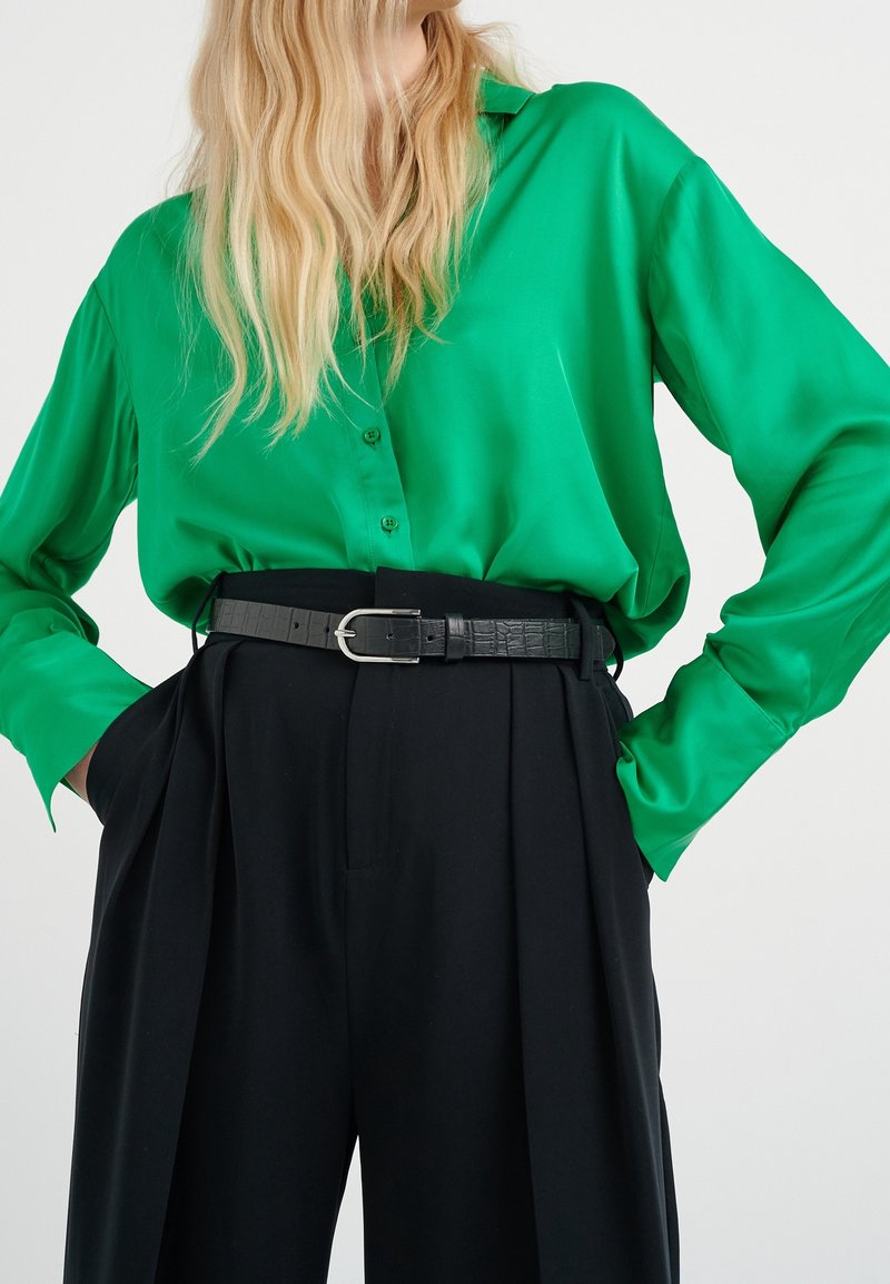 Blusa in raso verde con bottoni sul davanti e maniche lunghe. Abbinata a pantaloni neri plissettati e una cintura nera texturizzata con fibbia argentata.