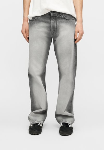 Άτομο που φορά ξεθωριασμένο γκρι τζιν με ίσια γραμμή, λευκό μπλουζάκι και μαύρα αθλητικά παπούτσια με λευκές ραβδώσεις, στέκεται μπροστά σε απλό φωτεινό φόντο.