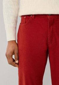 Pantalones de algodón rojos con una textura suave, con bolsillos laterales, cierre de botón en la parte delanteras y un ajuste Slim. Combinados con un suéter de punto en color crema.