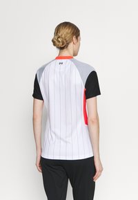 Koszulka z krótkim rękawem w kolorze białym z pionowymi czarnymi paskami, szarymi panelami na ramionach i czerwonymi akcentami. Wykonana z gładkiej, lekkiej tkaniny.