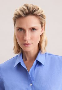 Camicia azzurro chiaro con bottoni, colletto e maniche corte, caratterizzata da una trama sottile. Il tessuto appare liscio e leggermente riflettente.