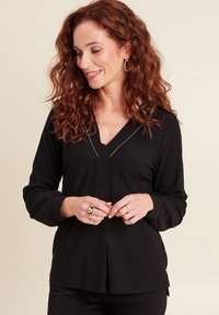 Haut noir à manches longues avec un décolleté en V, en tissu texturé, et un subtil accent argenté le long de l'encolure. Présente des fentes sur les côtés à l'ourlet.