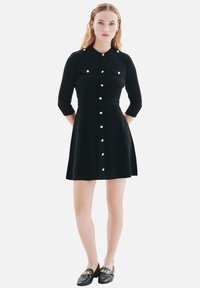Robe chemise noire avec un col, des manches trois-quarts, des boutons sur le devant et deux poches poitrine ; fabriquée en tissu lisse, coupe ajustée.