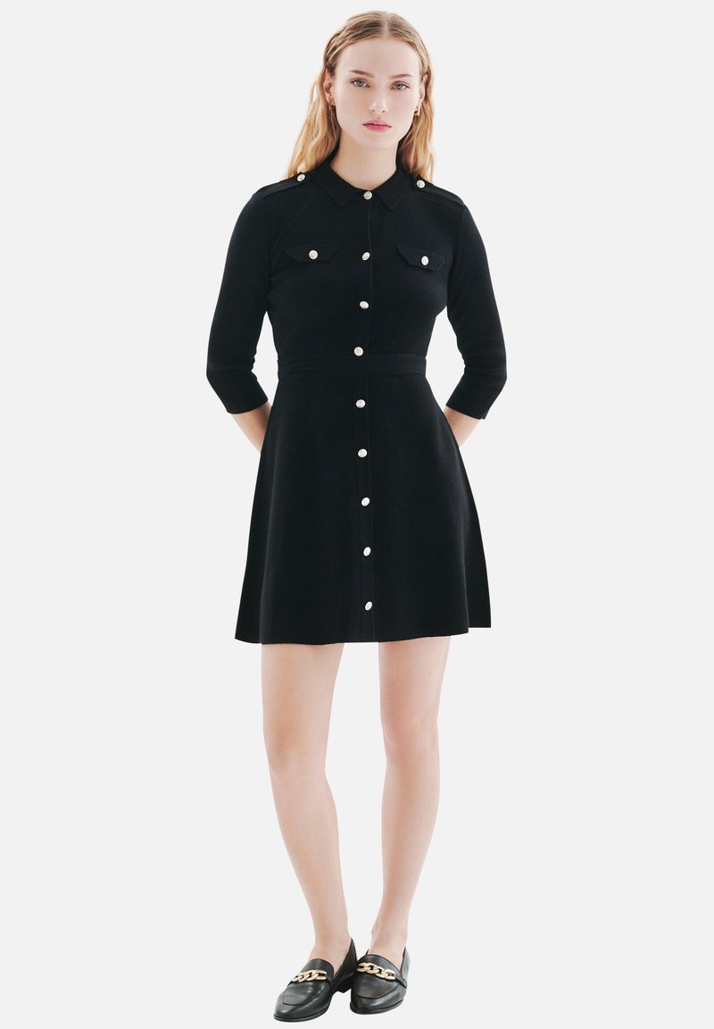 Robe chemise noire avec un col, des manches trois-quarts, des boutons sur le devant et deux poches poitrine ; fabriquée en tissu lisse, coupe ajustée.