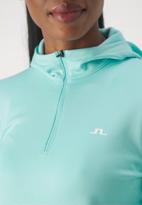 Světle tyrkysová mikina s půlzipem vyrobená z hladkého materiálu, se malým bílým logem a stahovací kapucí pro nastavitelné zakrytí.