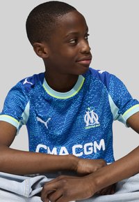 Dječak u plavoj majici s uzorkom Olympique de Marseille sjedi prekriženih nogu, gleda udesno s laganim osmijehom.