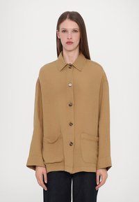 Tan oversized jacket with a collar, front button closure, and two front pockets. Features a textured fabric and wide sleeves.