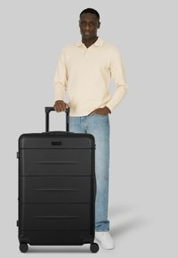 Valigia rigida nera con scanalature incassate, manico estensibile e quattro ruote. Su sfondo grigio neutro.