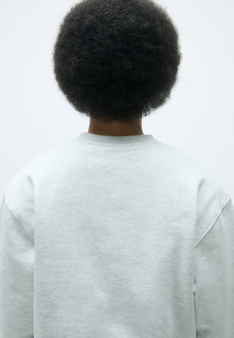 Hellgrauer Sweatshirt mit rundem Halsausschnitt und langen Ärmeln. Der Stoff wirkt weich und leicht strukturiert. Die Rückansicht zeigt eine lockere Passform.