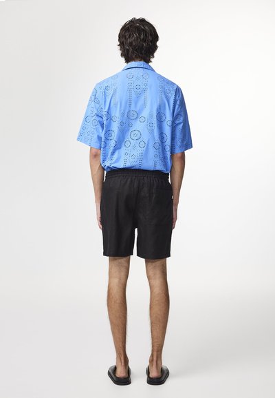Camicia azzurro chiaro a maniche corte con motivi circolari tagliati al laser, abbinata a pantaloni corti neri. Modello mostrato di spalle, con sandali neri.