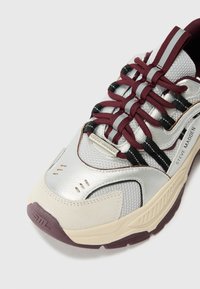 Sportschoenen met een zilverkleurige synthetische bovenkant, maroon en zwarte veters, beige accenten en een gestructureerde zool met merkdetails.