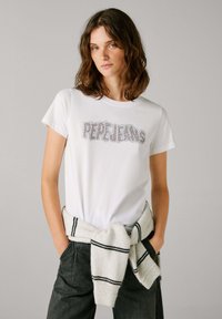 Camiseta de algodón blanca con "PEPE JEANS" en relieve de strass. Combinada con pantalones gris oscuro y un suéter crema atado a la cintura.