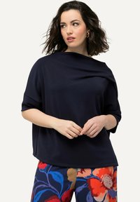 Top blu navy con colletto drappeggiato e maniche corte, abbinato a pantaloni a fantasia floreale vivace in rosso, blu e crema.