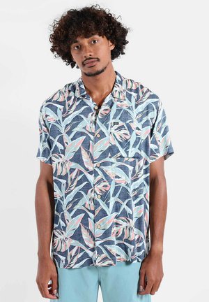 Homme aux cheveux bouclés portant une chemise boutonnée à manches courtes avec des motifs de feuilles tropicales bleu pastel et rose, et un pantalon bleu clair.