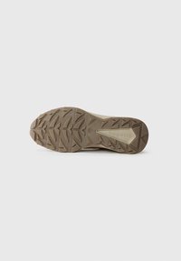 Semelle de chaussure avec une surface en caoutchouc texturé, présentant un motif de traction robuste en brun clair, mis en valeur par un logo central et des contours angulaires.