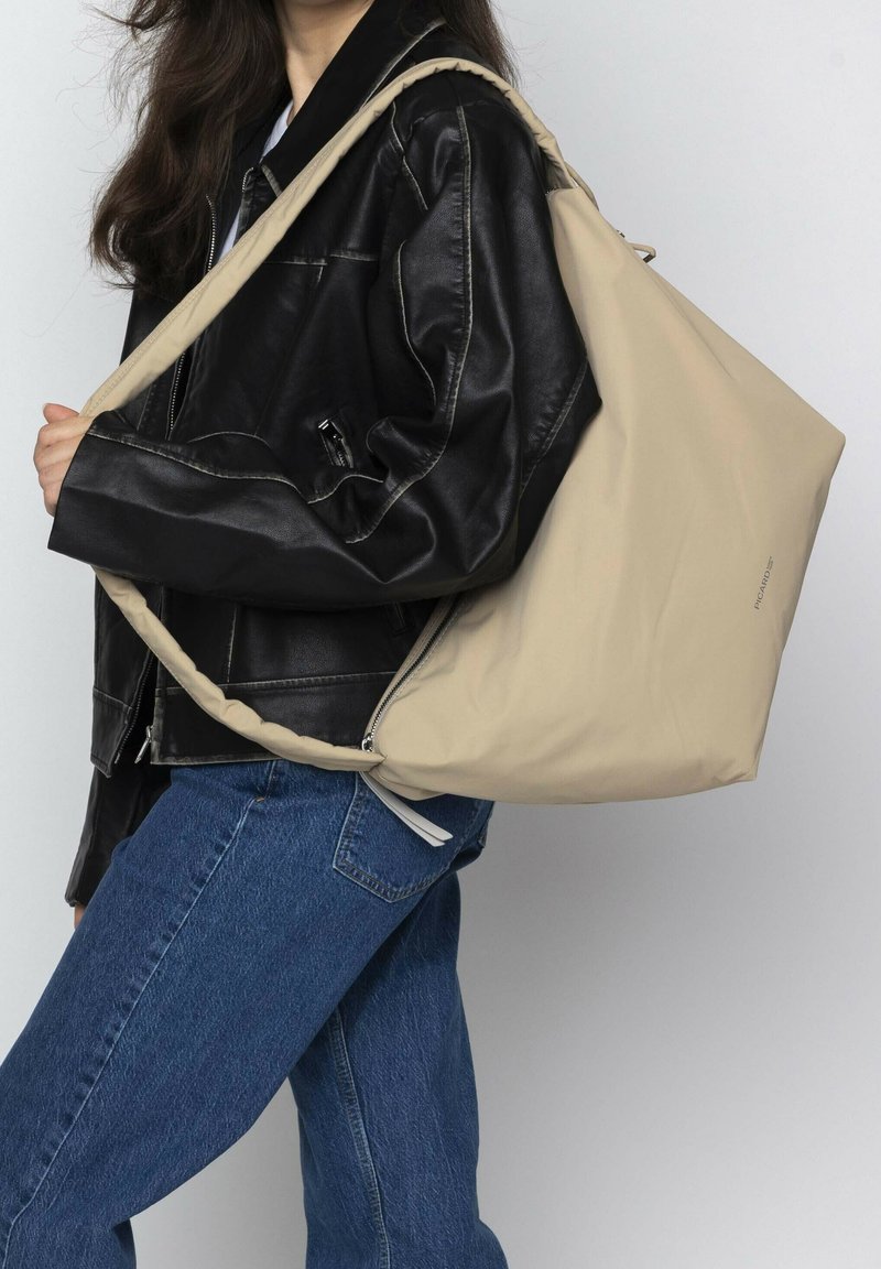 Bolso de nylon beige con una textura suave, diseño de gran tamaño y asa de hombro; combinado con una chaqueta de cuero negra y jeans azules.